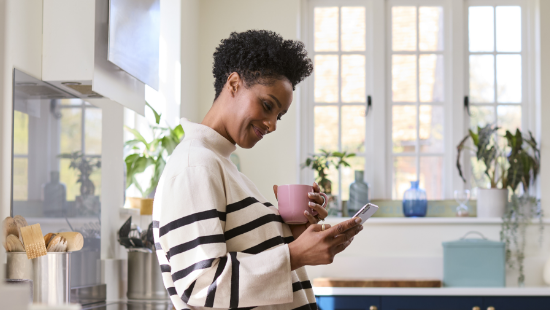 Young woman of color with short black hair is smiling and relaxing in her kitchen holding a mug looking at her cell phone